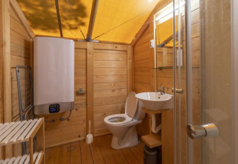 Bagno in tenda safari con pareti in legno, wc, lavandino, doccia, scaldabagno e pavimento in legno.