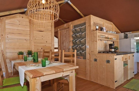 Intérieur de tente safari avec meubles en bois, table à manger, coin cuisine et décor vert.