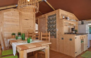 Interior of a safari tent with wooden furniture, dining table, compact kitchen, and green décor.