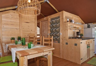 Interno di una tenda safari con mobili in legno, tavolo da pranzo, cucina e decorazioni verdi.