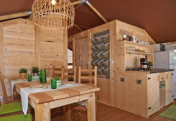 Interno di una tenda safari con mobili in legno, tavolo da pranzo, cucina e decorazioni verdi.