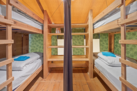 Interior de tienda safari con literas de madera, estantes, ropa de cama y toallas en cada cama.