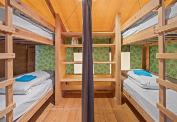 Binnenzicht van een safaritent met houten stapelbedden, opbergplanken en handdoeken op bedden.