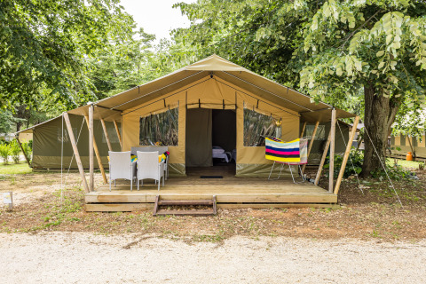 Tenda safari Harmony con veranda in legno, arredi da giardino e asciugamano colorato sotto gli alberi verdi.
