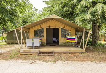 Tenda safari Harmony con veranda in legno, arredi da giardino e asciugamano colorato sotto gli alberi verdi.
