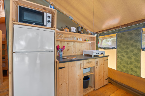 Coin cuisine de la Safaritent Relax avec micro-ondes, réfrigérateur, plaques, ustensiles et étagères en bois.