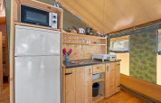 Kitchen area inside Safaritent Relax with microwave, fridge, hot plate, utensils, and wooden shelves inside tent.