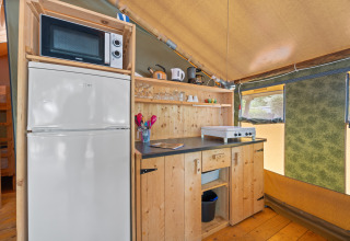 Coin cuisine de la Safaritent Relax avec micro-ondes, réfrigérateur, plaques, ustensiles et étagères en bois.