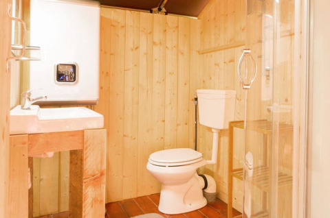 Badeværelse i safaritelts stil med træpaneler, håndvask, toilet og bruser med glasdør.