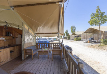 Veranda di una tenda safari con mobili in legno, cucina esterna e altre tende in un campeggio soleggiato.