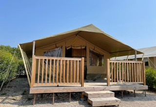 Tenda safari con ampia veranda in legno e corrimano, sotto un cielo azzurro in mezzo alla natura.