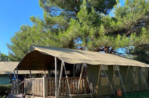 Tenda safari con veranda in legno tra alberi verdi sotto un cielo azzurro limpido in campeggio.