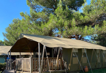 Tenda safari con veranda in legno tra alberi verdi sotto un cielo azzurro limpido in campeggio.