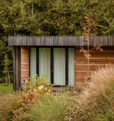 Photo de la Lodge, façade en bois moderne, grandes fenêtres et entourée de hautes herbes et de buissons.