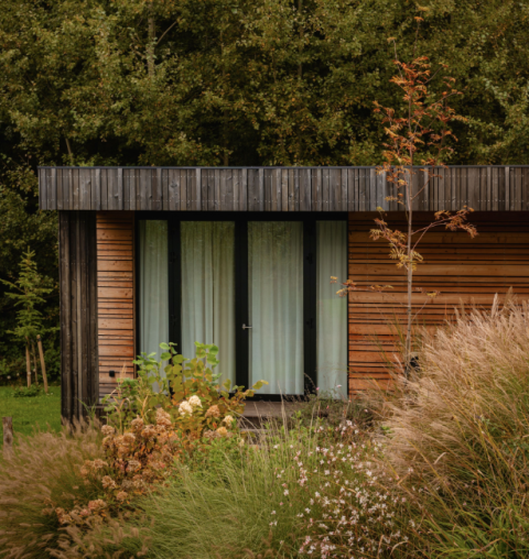 Eine Lodge mit moderner Holzfassade, großen Fenstern und umgeben von üppigen Büschen und Gräsern.