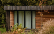 Photo de la Lodge, façade en bois moderne, grandes fenêtres et entourée de hautes herbes et de buissons.