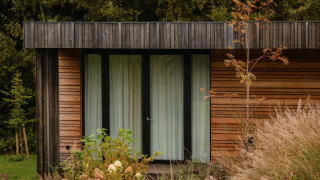 Photo de la Lodge, façade en bois moderne, grandes fenêtres et entourée de hautes herbes et de buissons.