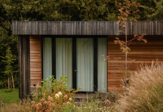 Photo de la Lodge, façade en bois moderne, grandes fenêtres et entourée de hautes herbes et de buissons.