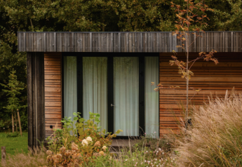 Photo de la Lodge, façade en bois moderne, grandes fenêtres et entourée de hautes herbes et de buissons.
