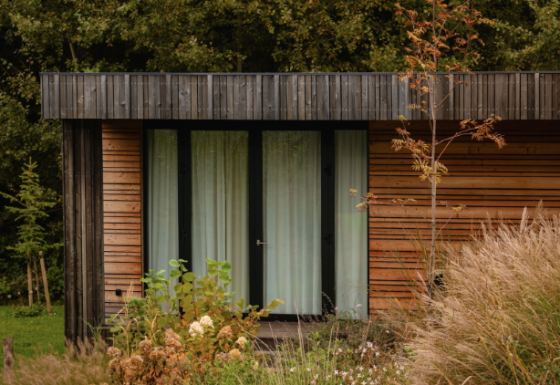 Photo de la Lodge, façade en bois moderne, grandes fenêtres et entourée de hautes herbes et de buissons.