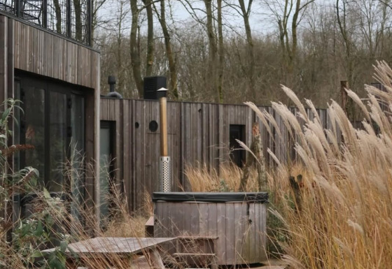 Houten terras met picknicktafels en hottub tussen hoog gras bij moderne lodge in Resort de Parel, Nederland.
