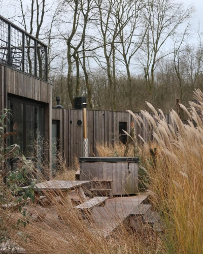 Houten terras met picknicktafels en hottub tussen hoog gras bij moderne lodge in Resort de Parel, Nederland.