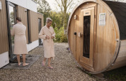 Two people in bathrobes at a lodge, one walking towards a barrel sauna, the other entering the main building.
