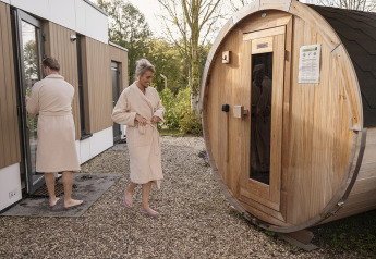 Due persone in accappatoio presso un lodge, una si avvicina alla sauna a botte, l'altra entra nell'edificio.