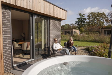 Modern lodge con due persone che si rilassano all’aperto vicino a una vasca idromassaggio a Hofparken Wiltershaar, Paesi Bassi.