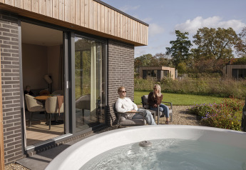Moderne Lodge mit zwei Personen, die sich draußen am Whirlpool bei Hofparken Wiltershaar, Niederlande entspannen.