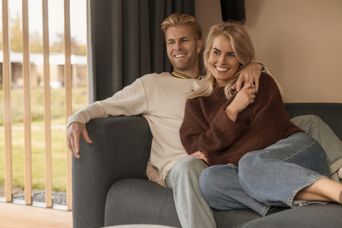 Un couple souriant est assis ensemble sur un canapé confortable dans un lodge, près d'une grande fenêtre.