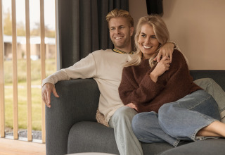 Una pareja sonriente sentada junta en un sofá acogedor de una cabaña, con grandes ventanales y vistas.