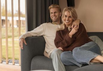 Una pareja sonriente sentada junta en un sofá acogedor de una cabaña, con grandes ventanales y vistas.