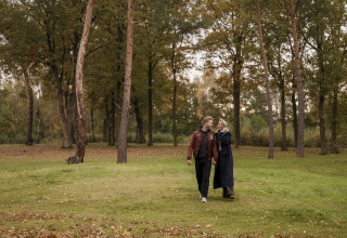 A couple walks hand in hand on a green lawn near a lodge, surrounded by tall autumn trees in the background.