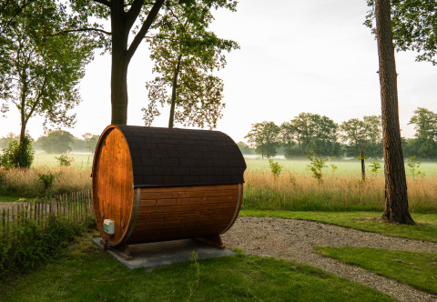 Een tonvormige sauna uit hout staat op het gras aan een pad, met bomen en een veld op de achtergrond.