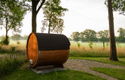 Eine fassförmige Holzsauna steht auf einer Wiese im Grünen, umgeben von Bäumen und einem Kiesweg.