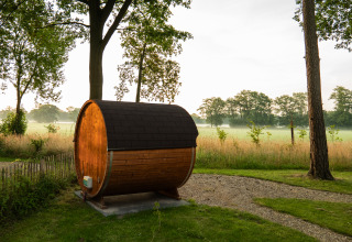 A barrel-shaped wooden sauna sits on a grassy area beside a path, with trees and fields in the background.