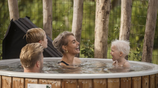 En smilende familie nyder en varm udendørs badetønde omgivet af træer og natur ved en lodge.