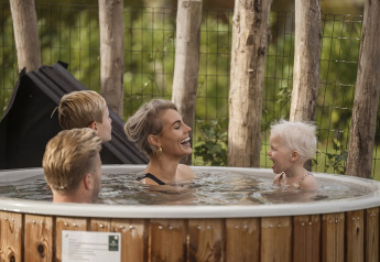 Een glimlachend gezin ontspant in een houten hottub buiten, omgeven door natuur aan een lodge.