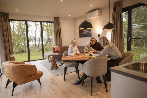 Familie spiller brætspil ved spisebordet i en hyggelig lodge med store vinduer og en hund på tæppet.