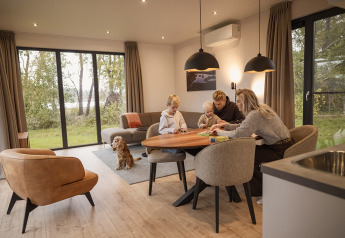 Familie spiller brætspil ved spisebordet i en hyggelig lodge med store vinduer og en hund på tæppet.