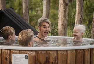 Une famille joyeuse profite d’un bain nordique extérieur en bois dans un lodge entouré de verdure.