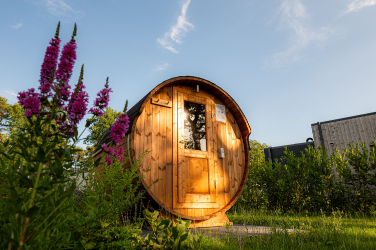 Sauna de madera con forma de barril en Holiday villa Amalia 2, rodeada de flores y vegetación en Hofparken Wiltershaar.