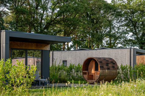 Lodge moderne avec grandes baies vitrées et sauna tonneau, entouré de verdure et d’arbres.