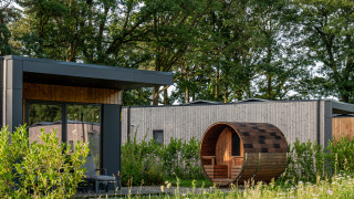 Moderne lodge med stort vindue og tøndeformet sauna i grønne omgivelser med træer og blomster.