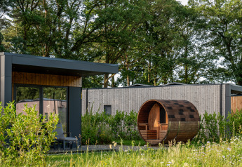 Modern lodge with large windows and barrel sauna surrounded by greenery and trees in countryside.