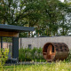 Moderne Lodge mit großer Glasfront und fassförmiger Sauna inmitten grüner Natur und Bäumen.