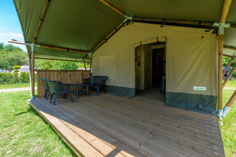Safari-Zelt mit überdachter Holzterrasse, Stühlen und Tisch, umgeben von grüner Natur im Freien.