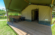 Safari-Zelt mit überdachter Holzterrasse, Stühlen und Tisch, umgeben von grüner Natur im Freien.