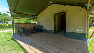 Tente safari avec terrasse en bois couverte, chaises et table, entourée d’herbe et de verdure.